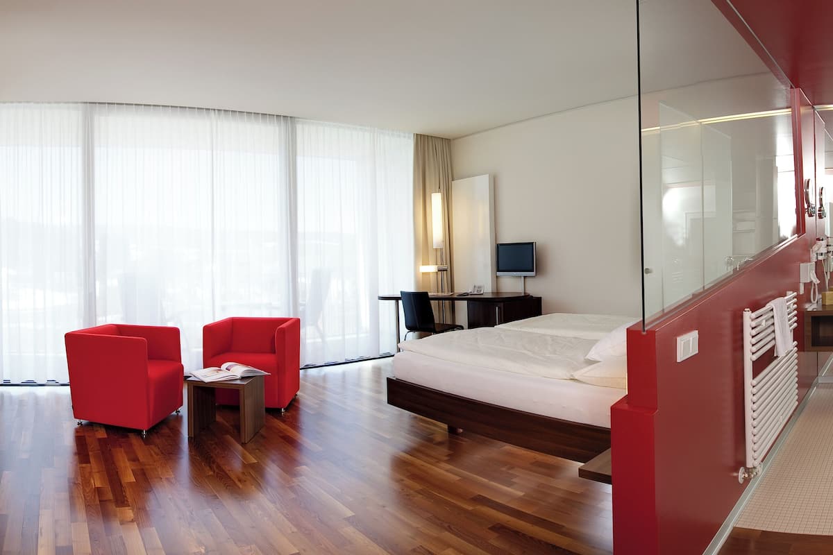 Moderni hotellihuone, jossa on suuri sänky, työpöytä tuolilla, televisio ja lasiseinä.
