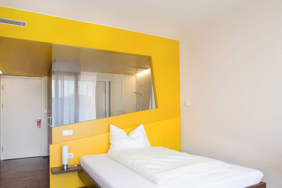 Moderni hotellihuone, jossa on puulattia, valkoisilla tekstiileillä varustettu sänky, yöpöytä lampun kanssa ja suuri peili seinällä.