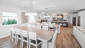 A modern kitchen with white cabinetry, a central island, and a dining area with a wooden table and white chairs. - Papa’s Beach House | 11BR/11.5BA Gulf Shores | Private Pool Coastal Escape (Gulf Shores)