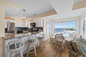A modern kitchen with white cabinets, a stainless steel refrigerator, and a black backsplash. There is a dining area with white wicker chairs and a patterned sofa.