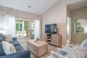 A living room with a sofa, coffee table, TV, and a sliding glass door leading to an outdoor area. - TB 81C Updated and spacious condo in True Blue with pool access (Pawleys Island)