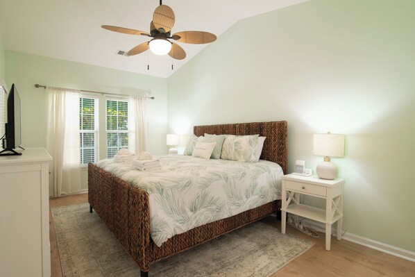 Une chambre à coucher avec un lit, une table de chevet avec une lampe, un ventilateur de plafond et une fenêtre avec des rideaux.