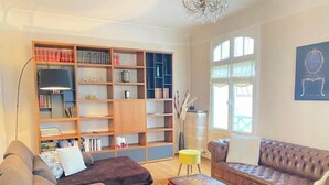 Un salón con un sofá marrón, un sillón marrón, una mesa de centro de madera y una amplia estantería llena de libros y objetos decorativos.