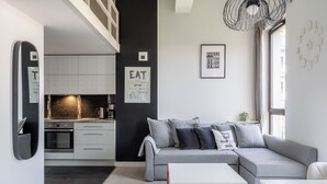 Un salon moderne avec un canapé gris, une table basse blanche et un coin cuisine doté de meubles blancs et d’un plan de travail noir.