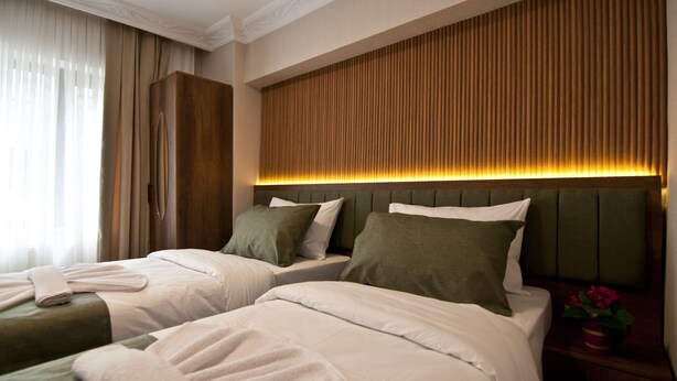 Ein Hotelzimmer mit zwei Betten, einem Holzkopfteil, einem Nachttisch mit Blumenvase und einem Fenster mit Vorhängen.