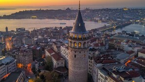 Eine Stadtansicht mit einem markanten Turm, der ein kegelförmiges Dach und einen Balkon hat, umgeben von dicht bebauten Gebäuden und einem Fluss im Hintergrund.