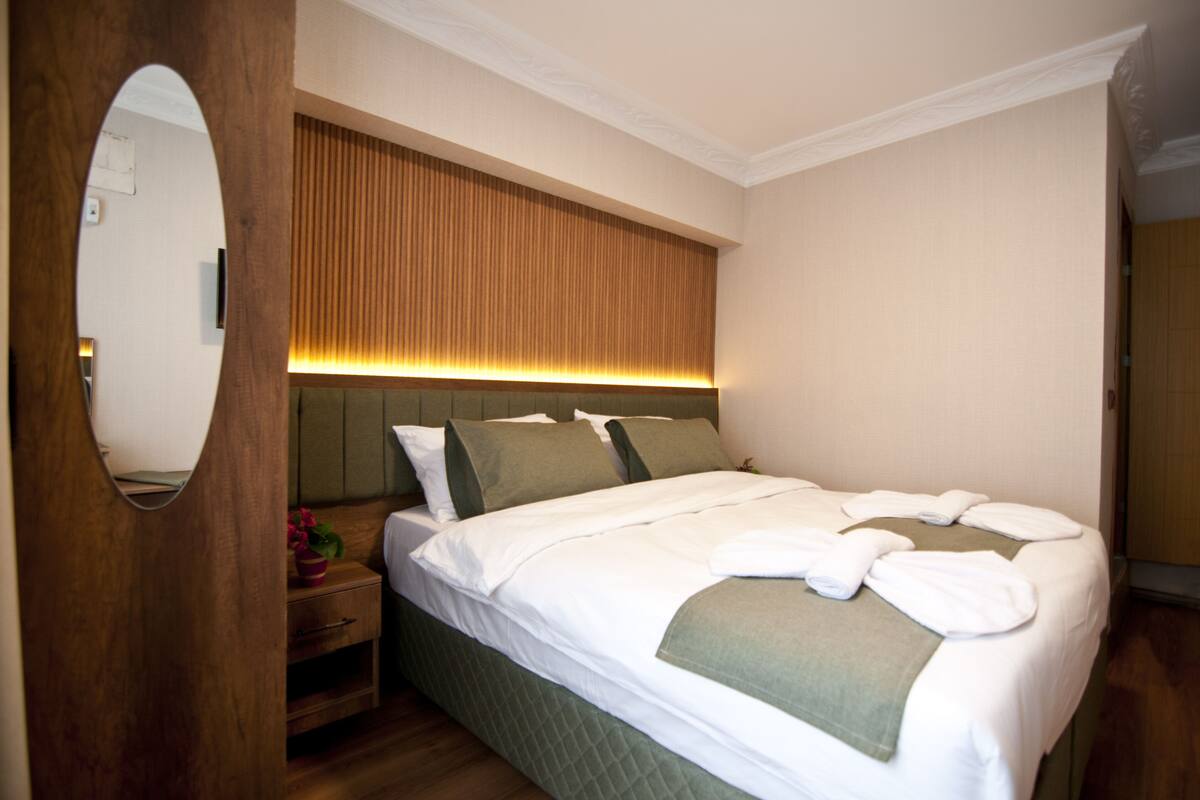 A neatly made bed with white linens and a green throw, a wooden nightstand with a pink flower vase, and a mirror on the left.