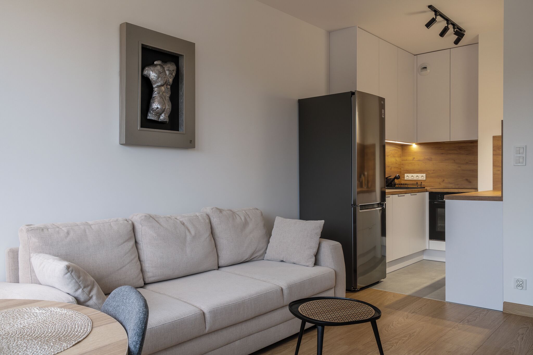 Una sala moderna con un sofá color beige, una mesita redonda y una zona de cocina con un refrigerador negro y armarios de madera.