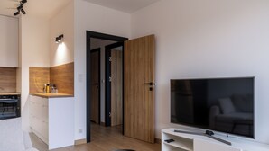 A modern living room with a large flat-screen TV, a sofa, a small round table, and a kitchen area with wooden cabinets and a countertop. - RentPlanet - Apartamenty Miedziana (Wroclaw)