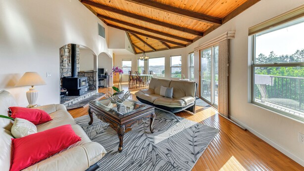A spacious living room with a high wooden ceiling, a stone fireplace, a sofa with red cushions, and a large window offering a view of the outdoors. - Kelseyville Home w/ Deck & Clear Lake Views! (Kelseyville)