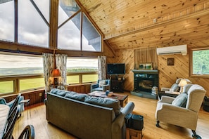 A spacious living room with a large window, a fireplace, a television, and a sofa. - Secluded Oxford Cabin on 75 Acres: Sunset Views! (Oxford)