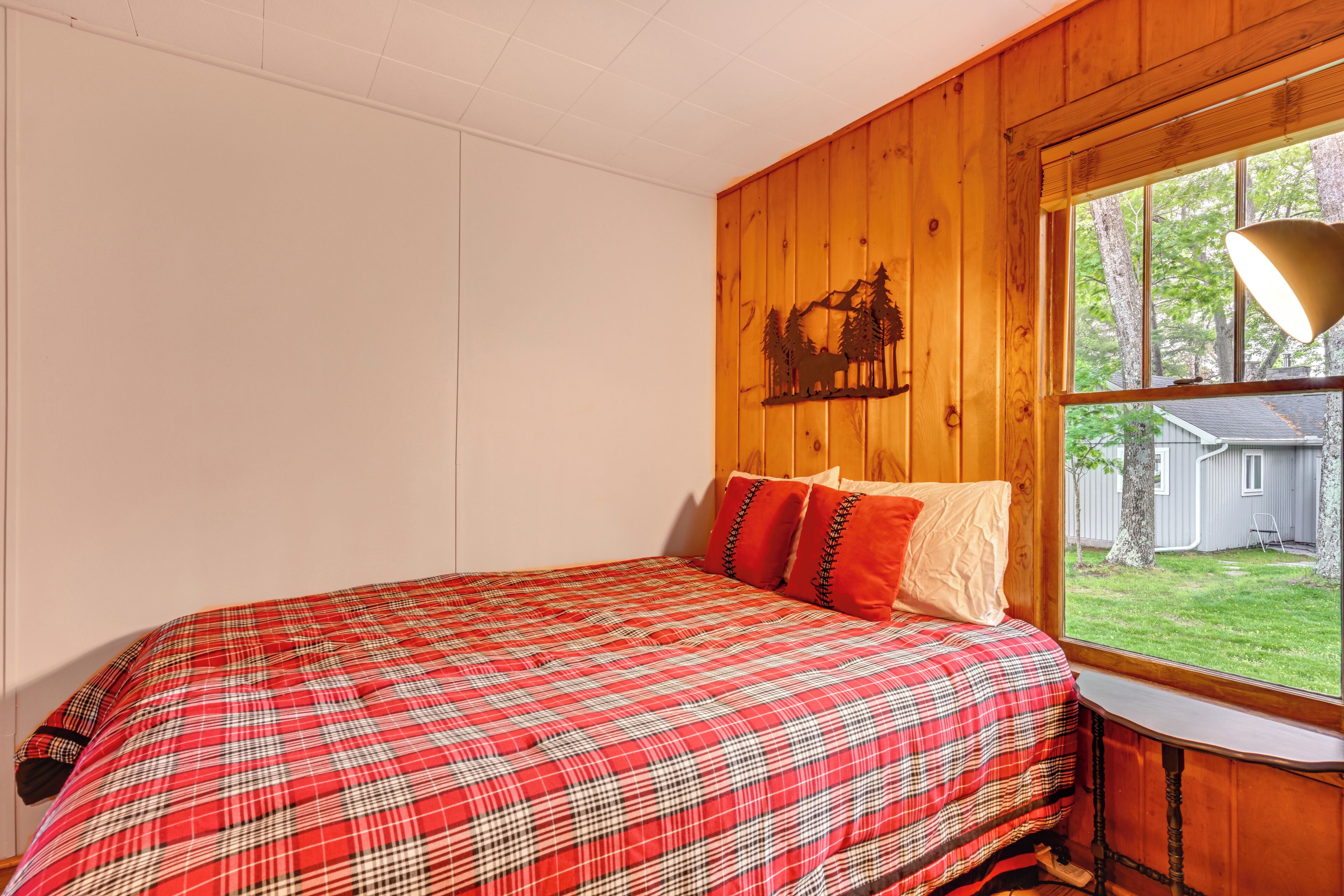A bedroom with a plaid bedspread, wooden walls, a window with a view of the outdoors, and a wall-mounted lamp.