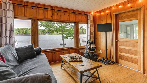Una habitación acogedora con detalles de madera, un ventanal con vistas a un lago, un sofá, una mesa con aperitivos y un televisor sobre un mueble.