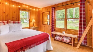 Un dormitorio de cabaña de madera con una cama, lámparas de noche, un banco con una manta escocesa y una ventana con cortinas.