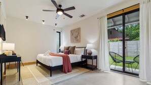 Un dormitorio moderno con cama, mesitas de noche, ventilador de techo y puerta corrediza de vidrio que da a un patio con mobiliario exterior.