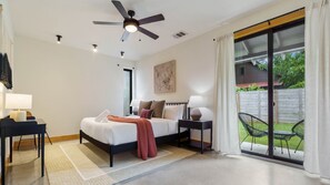 A modern bedroom with a bed, bedside tables, a ceiling fan, and a sliding glass door leading to a patio with outdoor furniture.