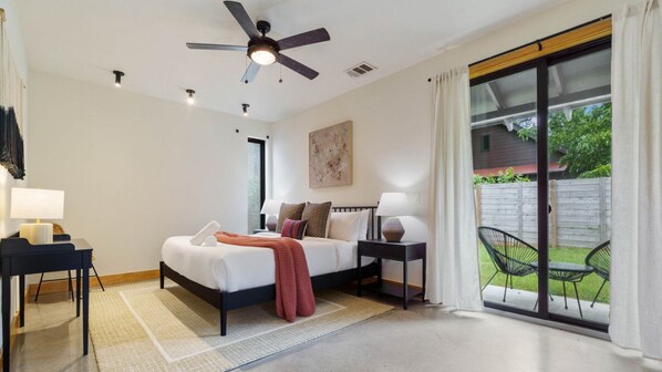 A modern bedroom with a bed, bedside tables, a ceiling fan, and a sliding glass door leading to a patio with outdoor furniture. - Caddie St #A xxxxxxx (Austin)