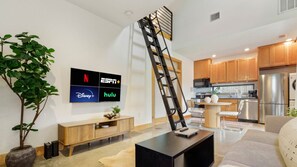 A modern living room with a TV displaying streaming services, a staircase, and a kitchen area. - Caddie St #A xxxxxxx (Austin)