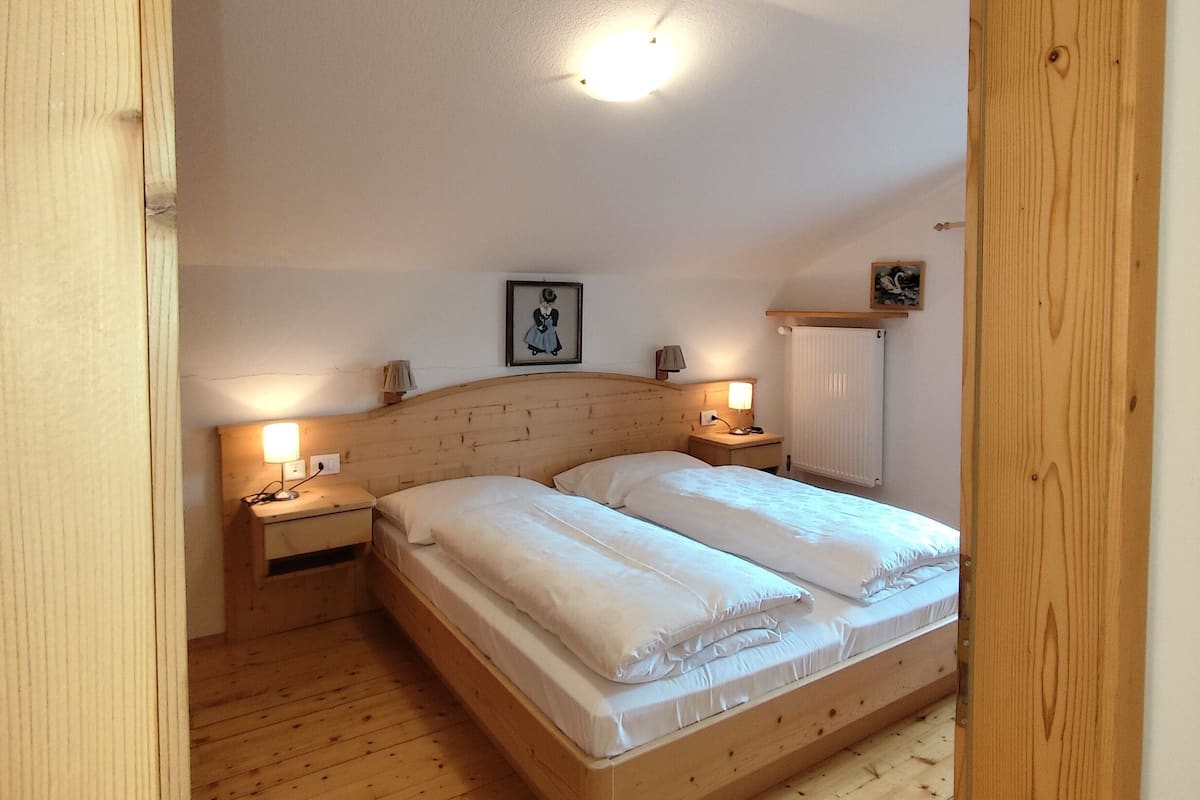 A bedroom with a wooden floor, a double bed, two bedside tables with lamps, a ceiling light, and a framed picture on the wall.
