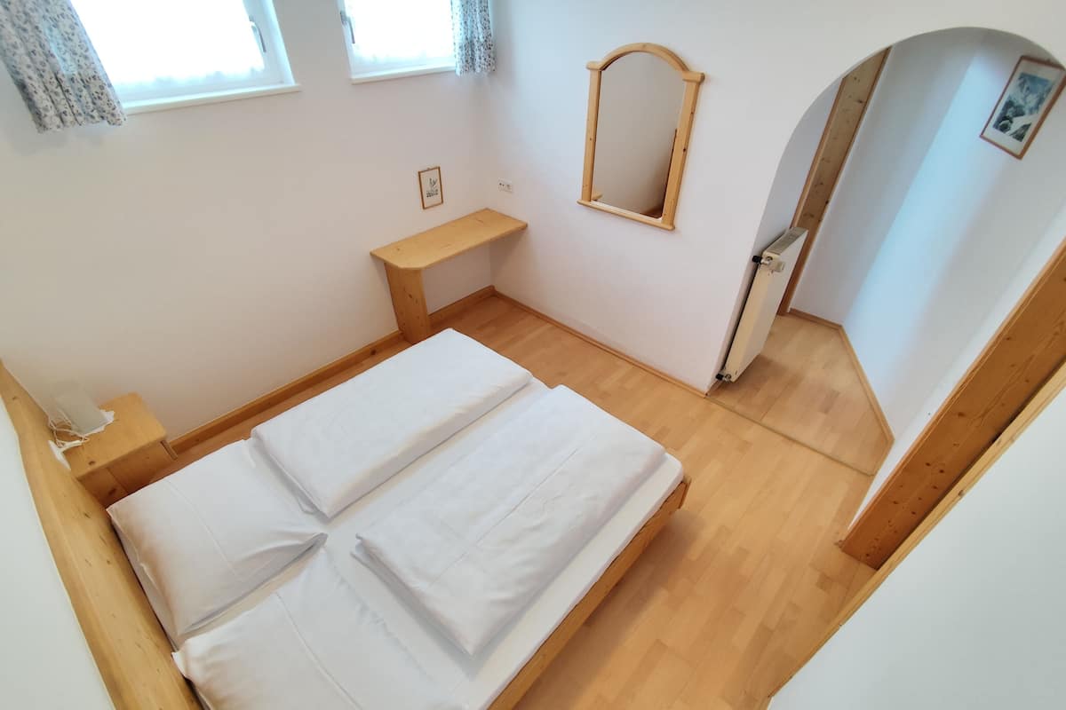 A bedroom with a wooden bed, a small bedside table, a mirror, and a window with curtains.