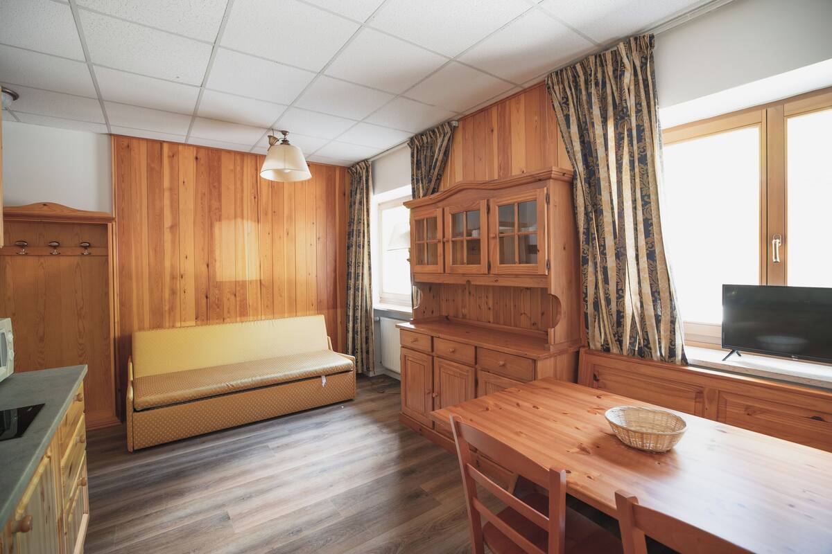 A room with wooden walls, a sofa, a dining table with chairs, a television, and a window with curtains.