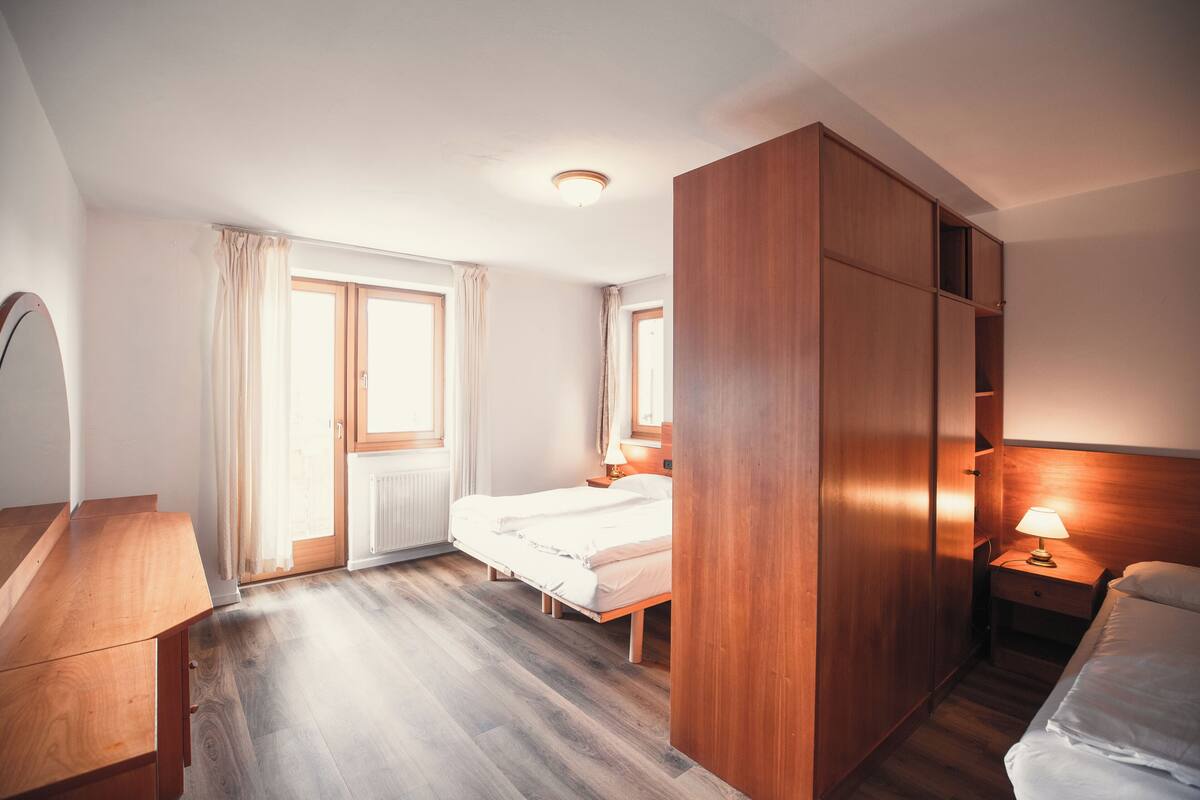 A bedroom with a wooden bed, a nightstand with a lamp, a wooden wardrobe, and a window with curtains.