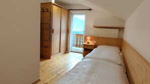Une chambre avec un plancher en bois, un lit avec une literie blanche, une armoire en bois, une petite table de chevet avec une lampe et une fenêtre donnant sur un balcon.