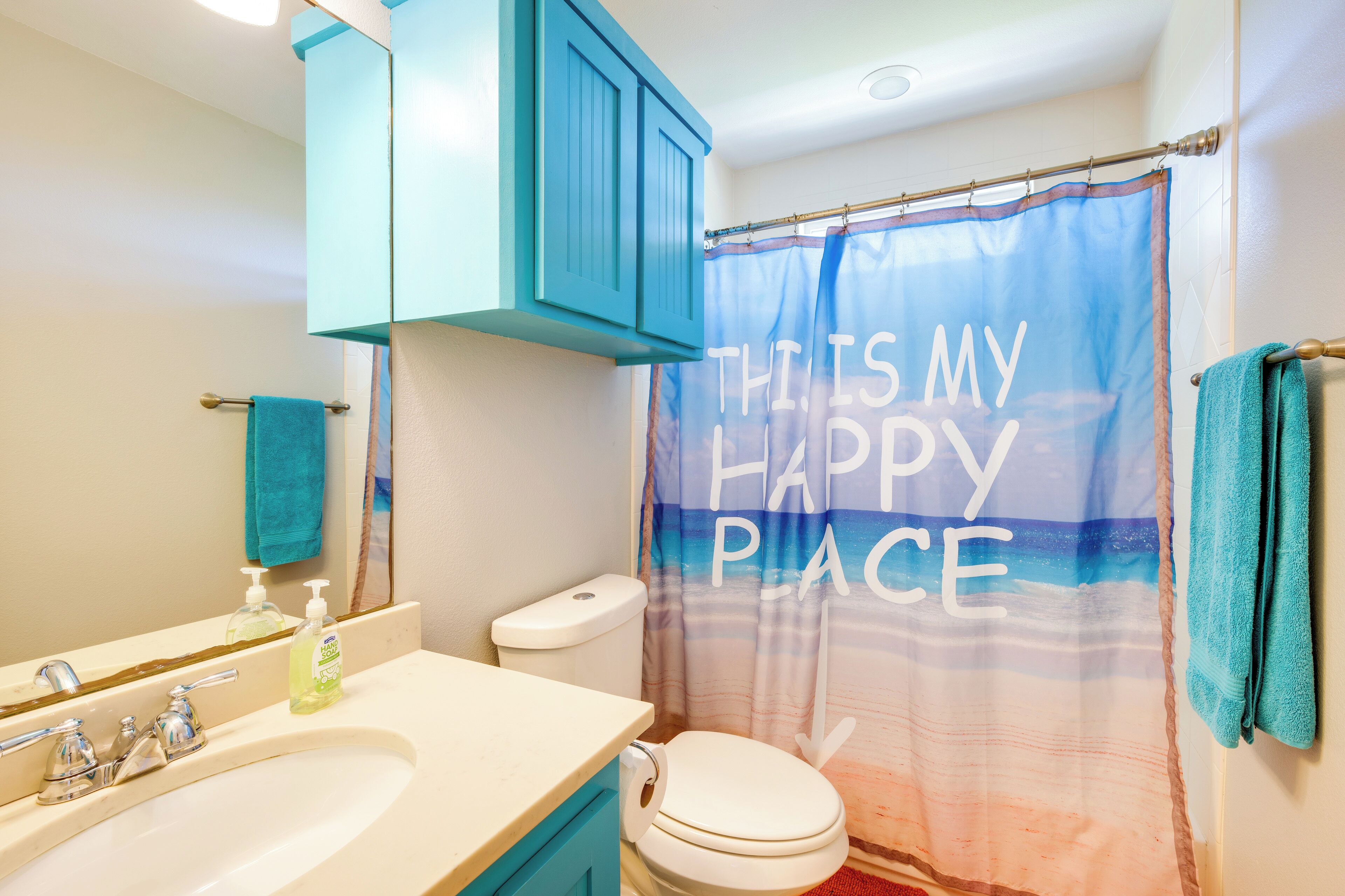 Une salle de bain avec un lavabo blanc, des toilettes et un rideau de douche sur lequel est écrit : « ICI, JE SUIS HEUREUX/HEUREUSE ».