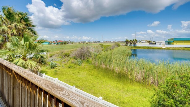 A view of a grassy area with a pond, a wooden deck, and buildings in the background. - Steps to Beach & Pool Access: Port Aransas Condo (Port Aransas)