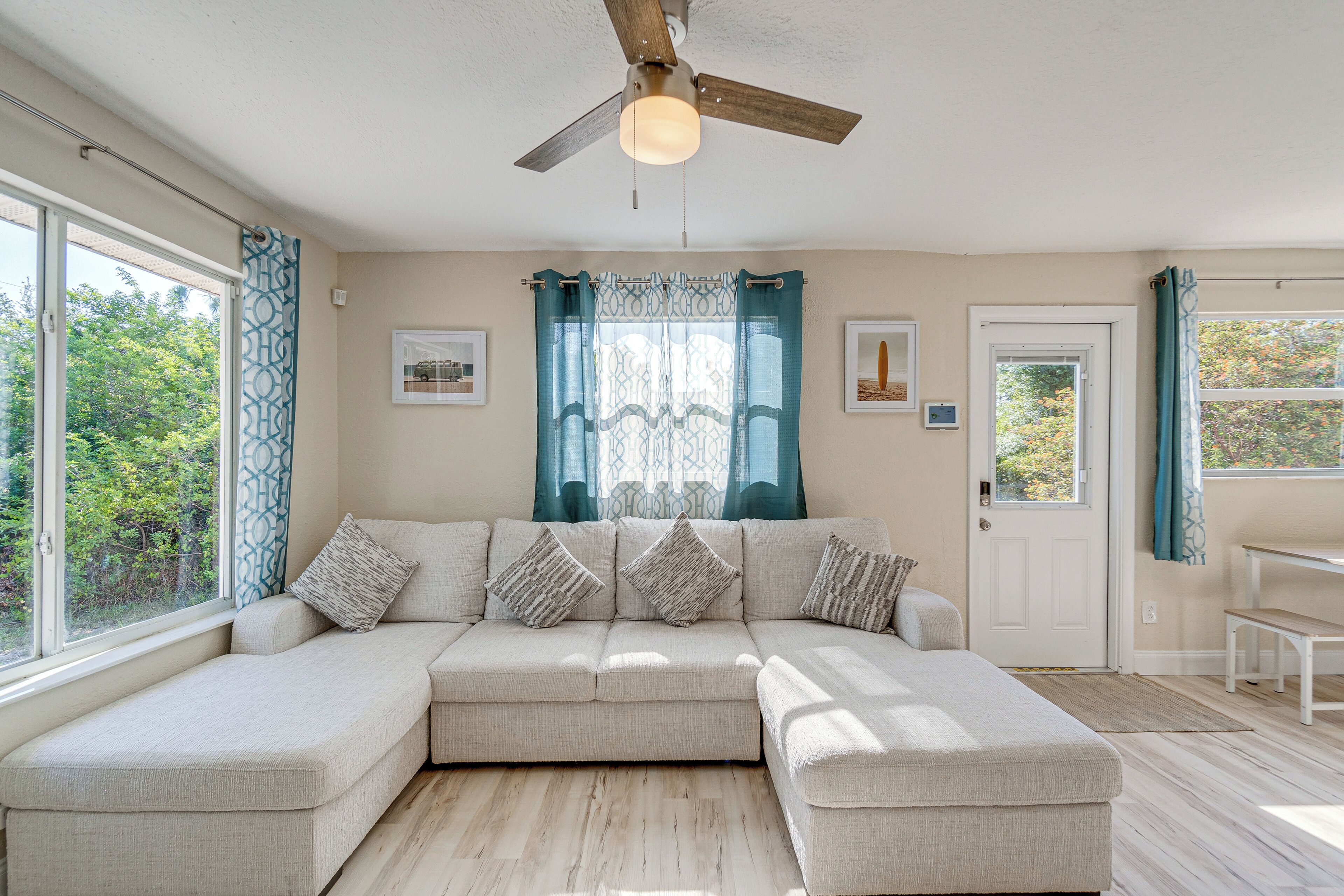 Un salon moderne avec un canapé d’angle, un ventilateur de plafond et de grandes fenêtres dotées de rideaux.