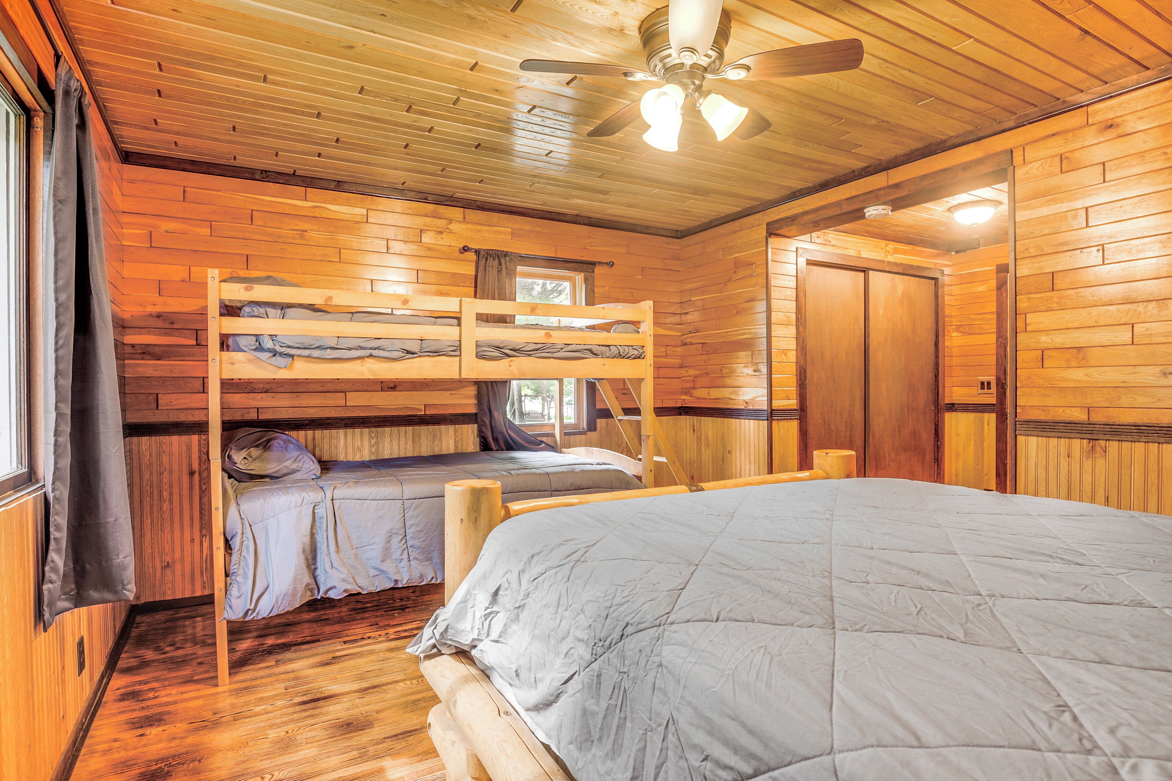 A cabin with a bunk bed, a single bed, wooden walls, a ceiling fan, and a window with curtains.