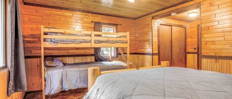 A cabin with a bunk bed, a single bed, wooden walls, a ceiling fan, and a window with curtains.