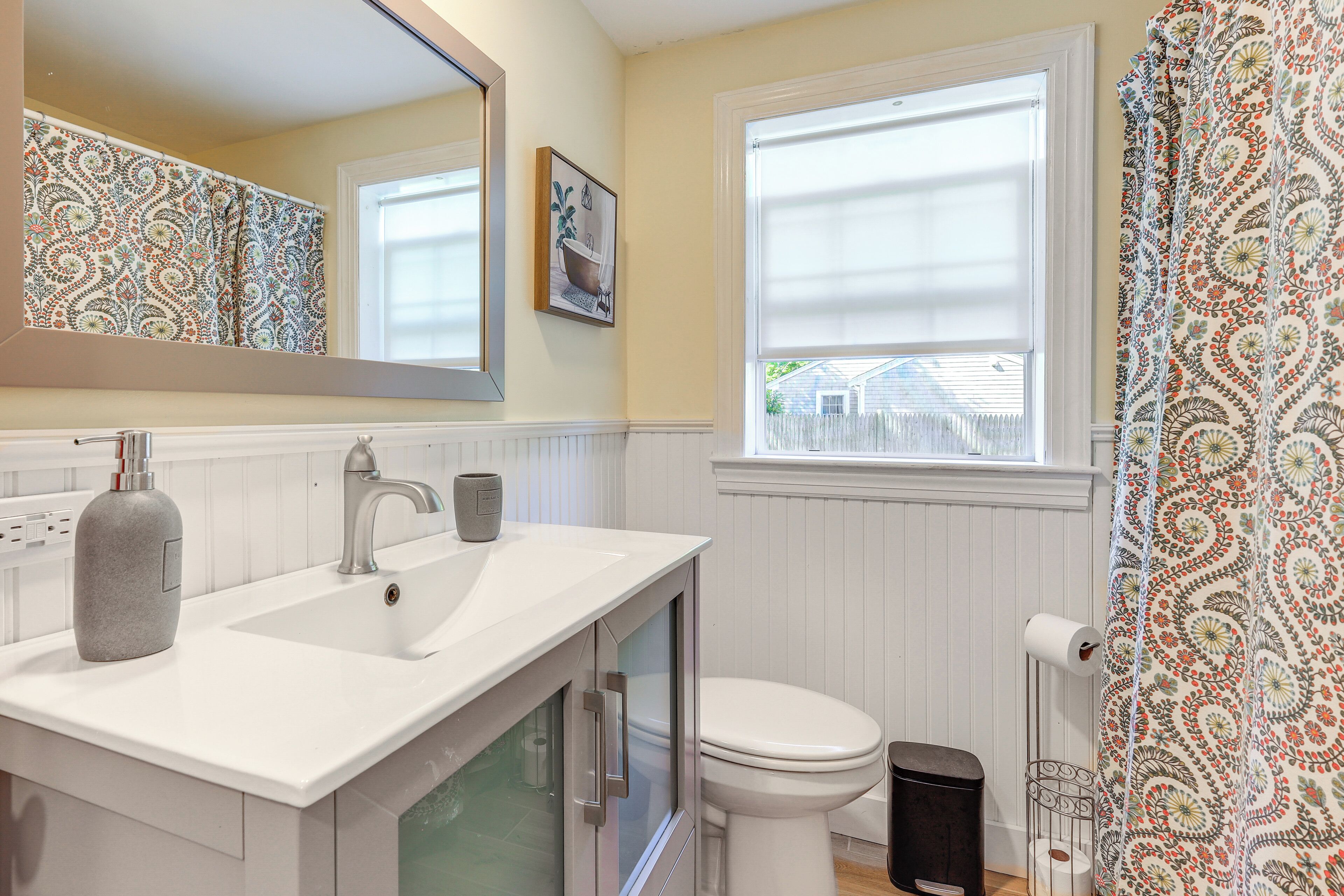 A bathroom with a white sink, a toilet, a window with blinds, and patterned curtains.