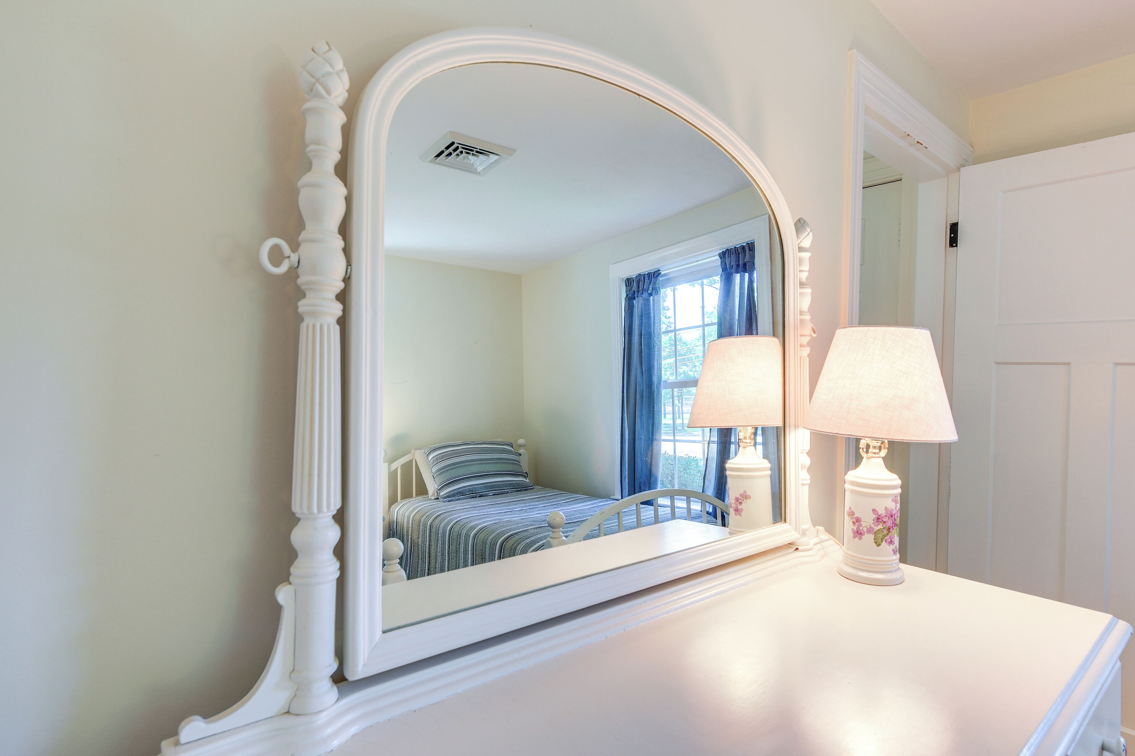 A bedroom with a large mirror, a bed with striped bedding, a bedside table with a lamp, and a window with blue curtains.