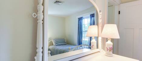 A bedroom with a large mirror, a bed with striped bedding, a bedside table with a lamp, and a window with blue curtains.