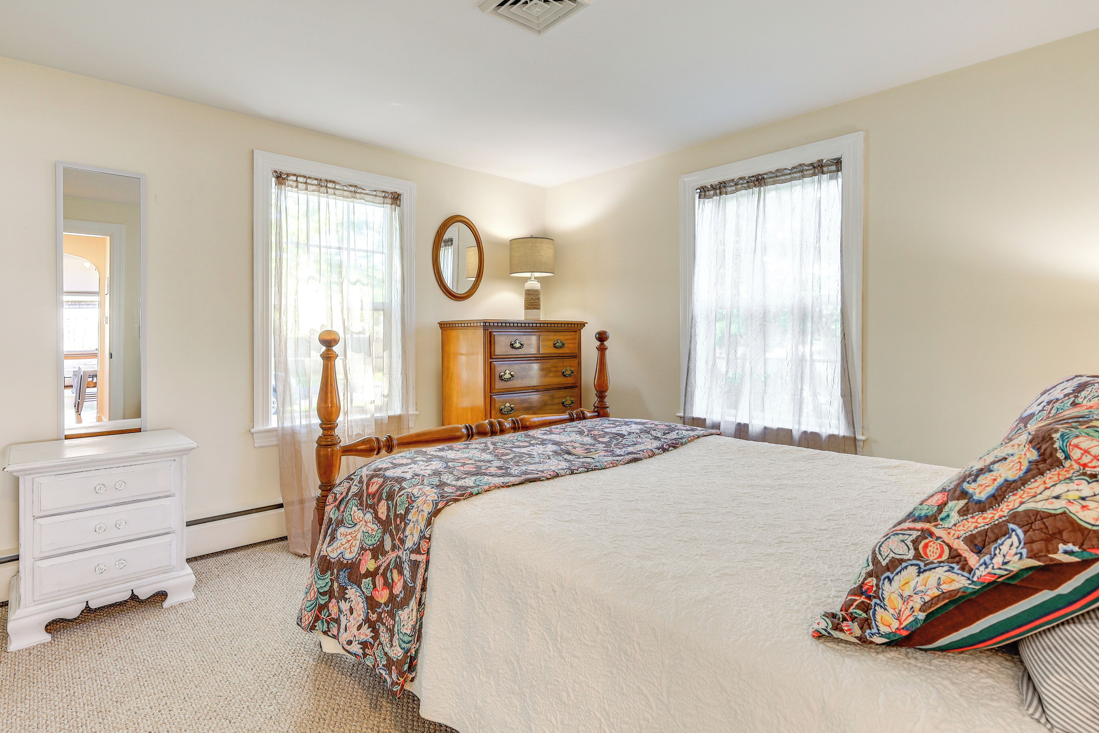 Une chambre à coucher comprenant un lit, une commode, un miroir et deux fenêtres avec des rideaux.