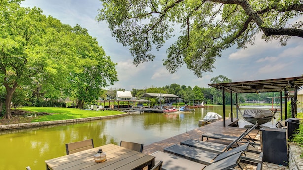 A wooden deck with tables and chairs overlooking a calm river, surrounded by lush greenery and a gazebo.
