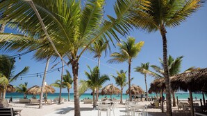 Zona junto a la playa con palmeras, sombrillas de paja y asientos al aire libre.