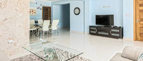 A modern living room with a glass coffee table, a beige sofa, a dining area with chairs, and a TV on a black cabinet.