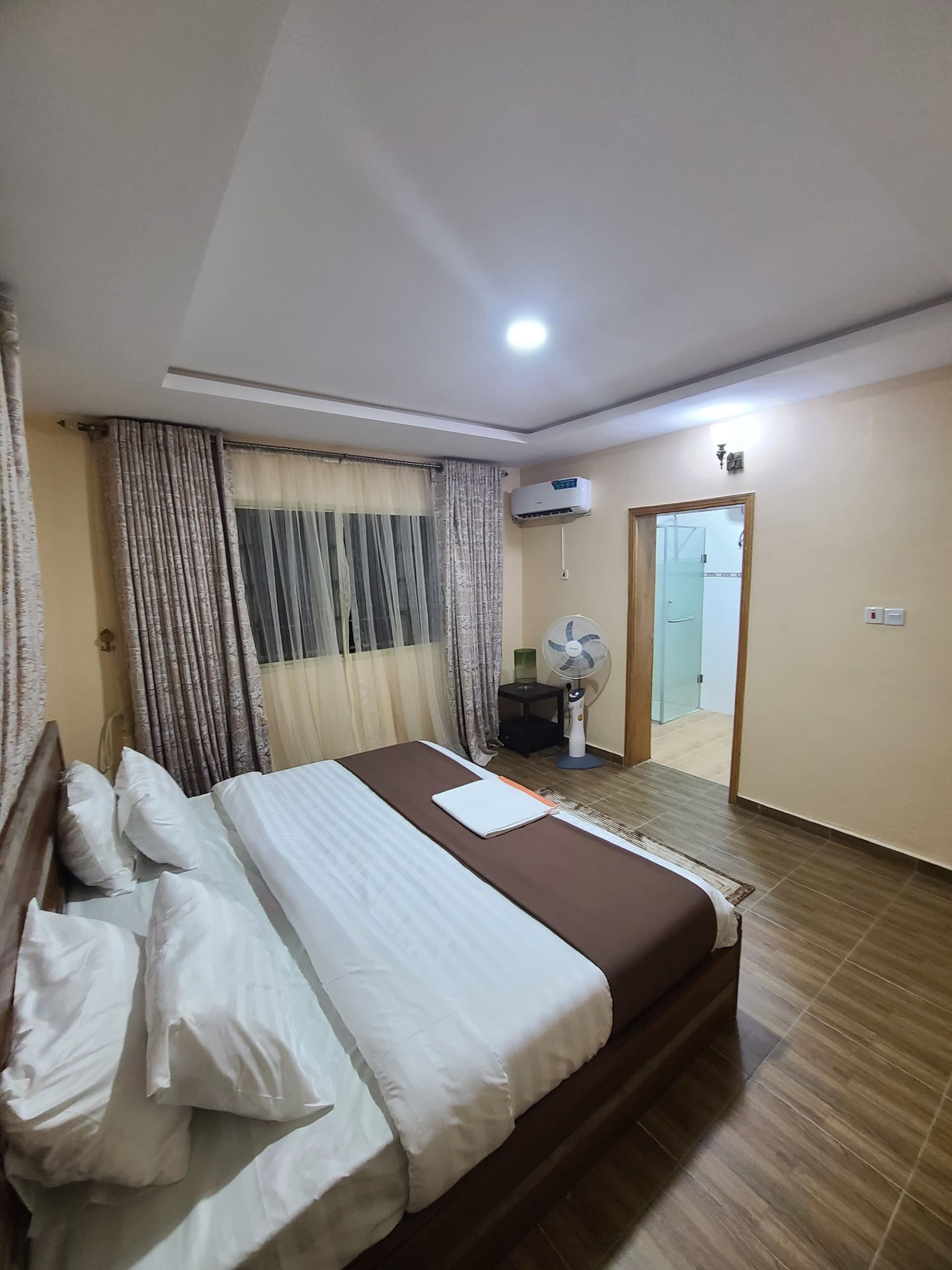 A hotel room with a bed, curtains, an air conditioning unit, a fan, and a door leading to another room.