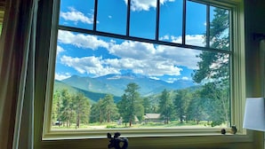 A room with a large window offering a view of a mountainous landscape with trees and a clear sky. - Best Views-Next to RMNP-Hike from House-HotTub-Fireplace-Coaster Passes-King Bed (Estes Park)