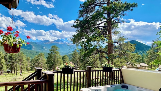 A wooden deck with a hot tub, surrounded by a wooden railing, overlooking a mountainous landscape with trees and a clear sky. - Best Views-Next to RMNP-Hike from House-HotTub-Fireplace-Coaster Passes-King Bed (Estes Park)