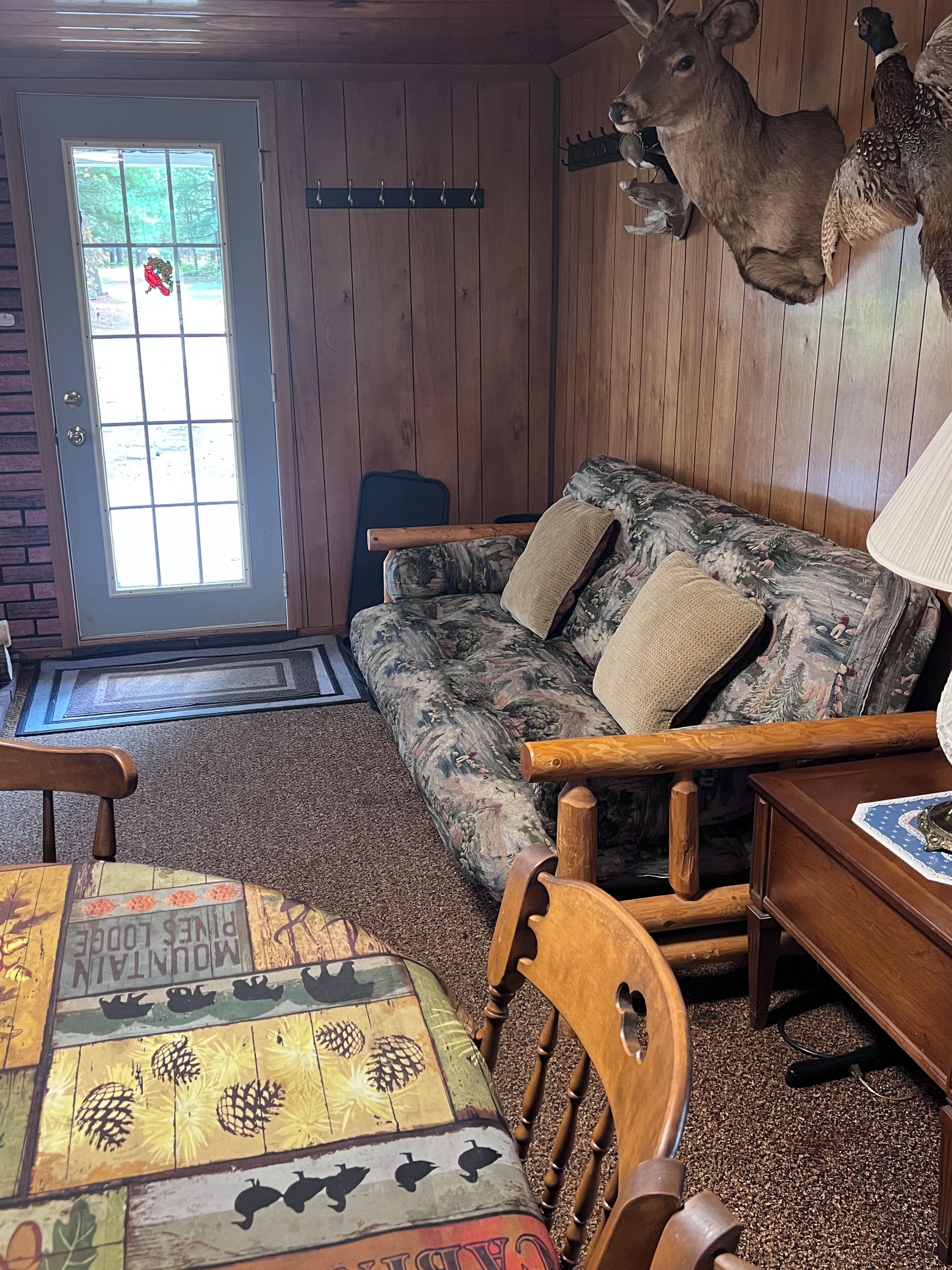 Interior acogedor de una cabaña con una puerta de madera, un sofá, una mesa de comedor con mantel estampado y una cabeza de venado montada en la pared.