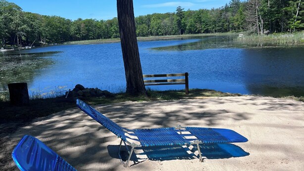 Une plage de sable avec deux chaises longues bleues et un banc en bois, près d’un lac.