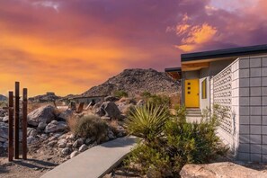 A modern building with a yellow door, surrounded by rocks and sparse vegetation, under a sunset sky.