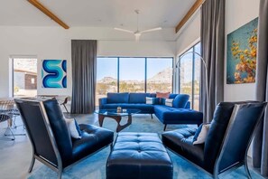 Un salon moderne avec un canapé d’angle bleu, des fauteuils en cuir noir et une table basse en verre. Une grande fenêtre offre une vue sur le paysage désertique.