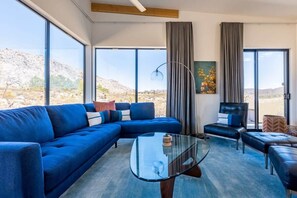 Una sala de estar moderna con un sofá seccional azul, una mesa de centro de vidrio y vistas a un paisaje desértico a través de amplias ventanas.