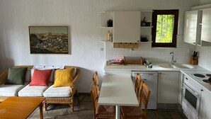 A compact kitchen with white cabinets, a built-in dishwasher, and a stove. There's a dining area with a white table and wicker chairs. A painting of a landscape hangs on the wall. - Maisonette With Garden Under the Elani Pines (Kassandra)