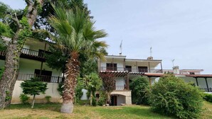 A two-story building with a balcony, surrounded by greenery and a palm tree. - Maisonette With Garden Under the Elani Pines (Kassandra)