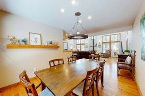 A spacious dining area with a wooden table and chairs, a chandelier, and a view of the living room through large windows. - Hot Tub, Dog Friendly, Great Location (Pagosa Springs)